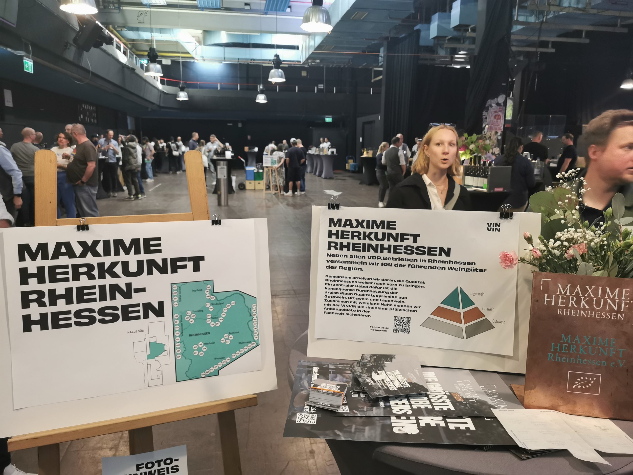 Indoor event hall with groups of people around display boards; foreground posters read 'MAXIME HERKUNFT RHEINHESSEN' beside a map UI element.