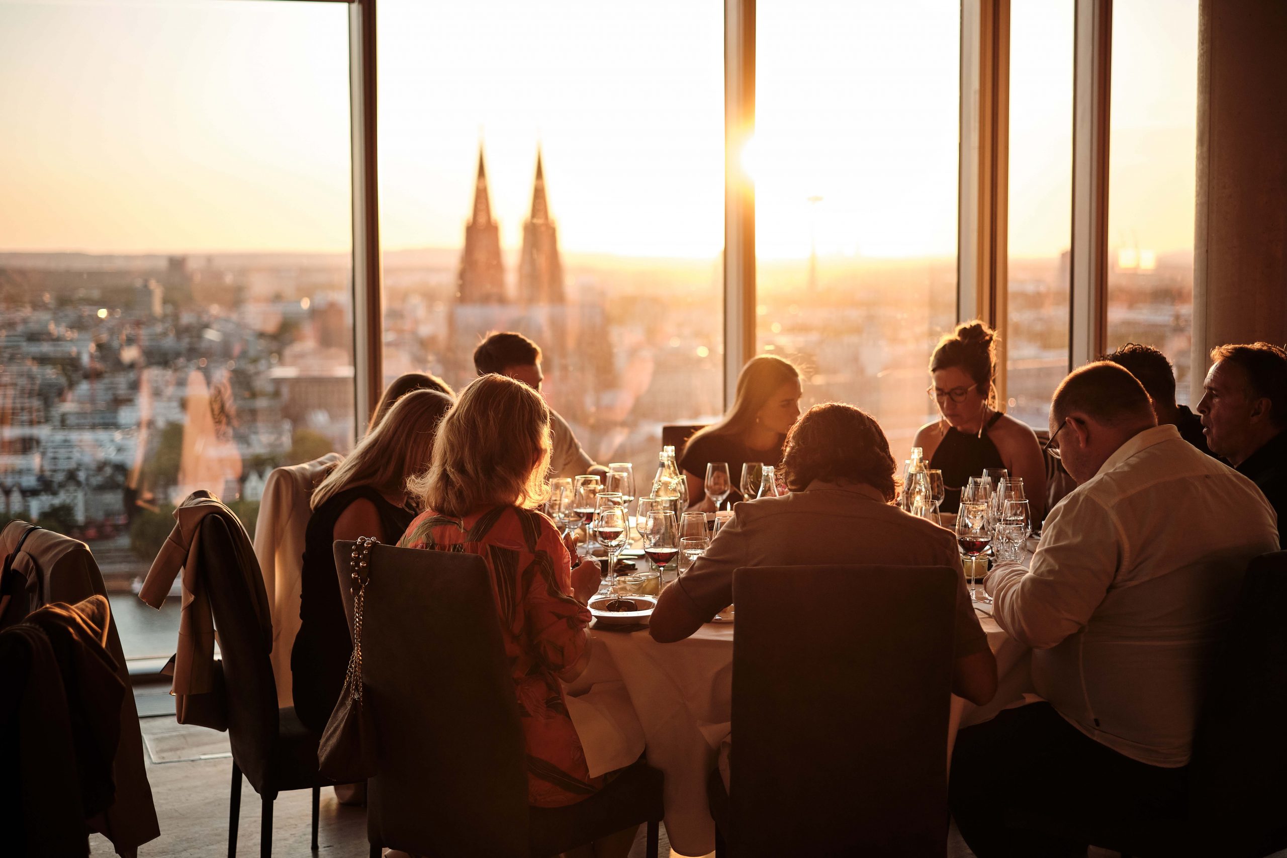 Group of people dining at a long table by large windows at sunset, with wine glasses and plates in front of them.