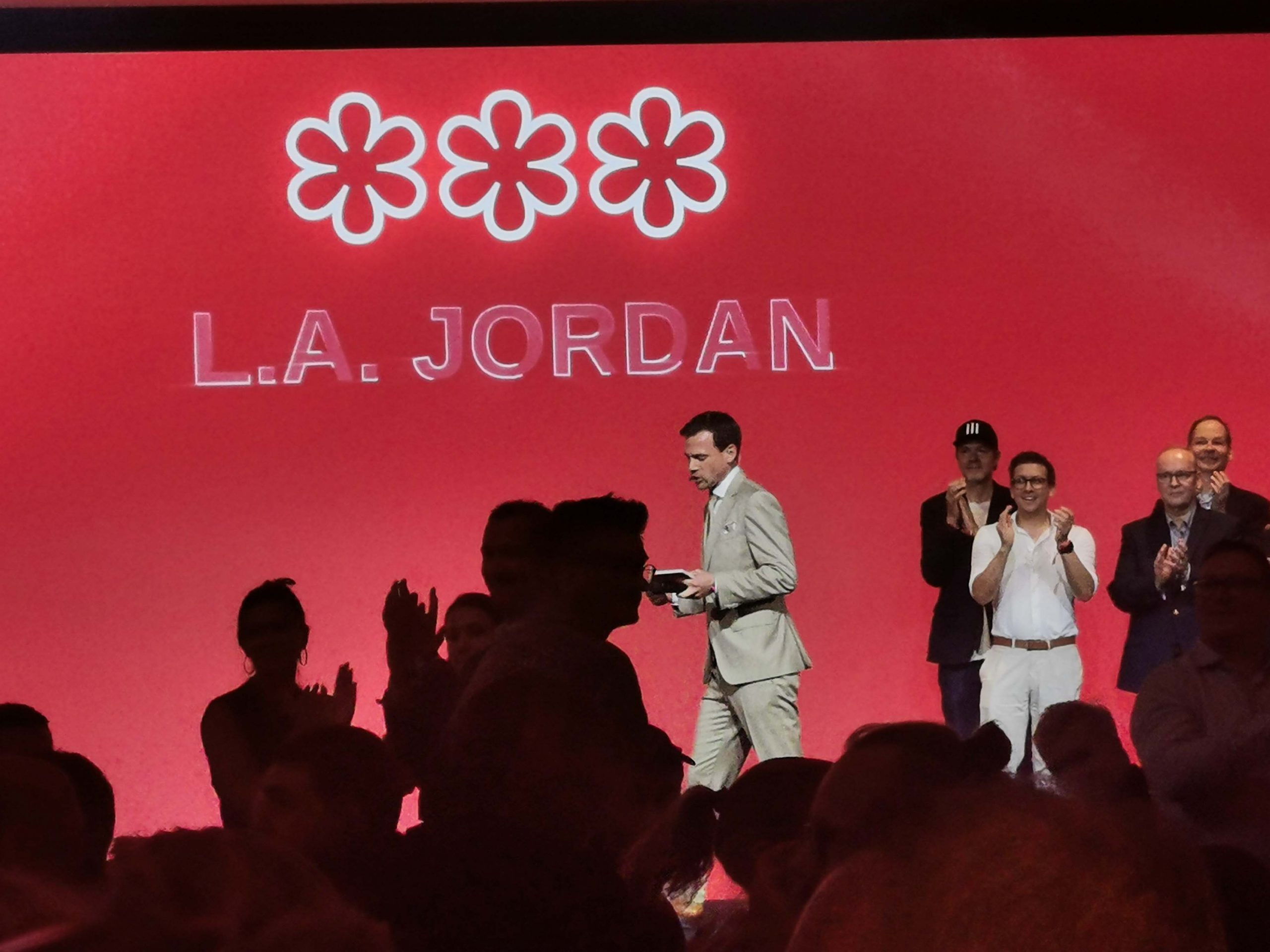 Group of people on a stage with a red backdrop, featuring three white flower logos and the text “L.A. JORDAN” in white; audience silhouettes in the foreground.