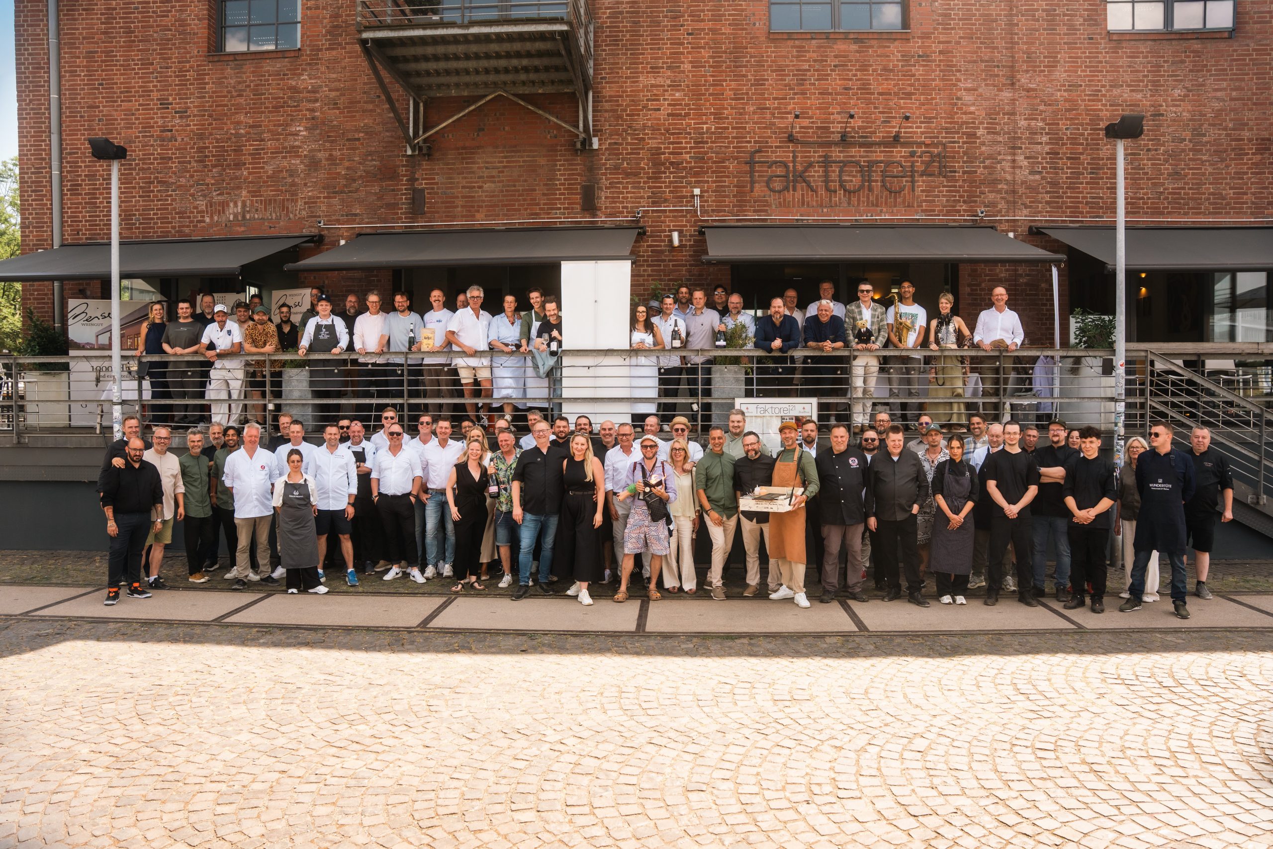 Pressefoto_Kulinaris~t_Tobias_Wacker_edited - ChefHeads-Magazin Large group of people posing on a brick building terrace outdoors for a group photo on a sunny day in front of a restaurant/venue.