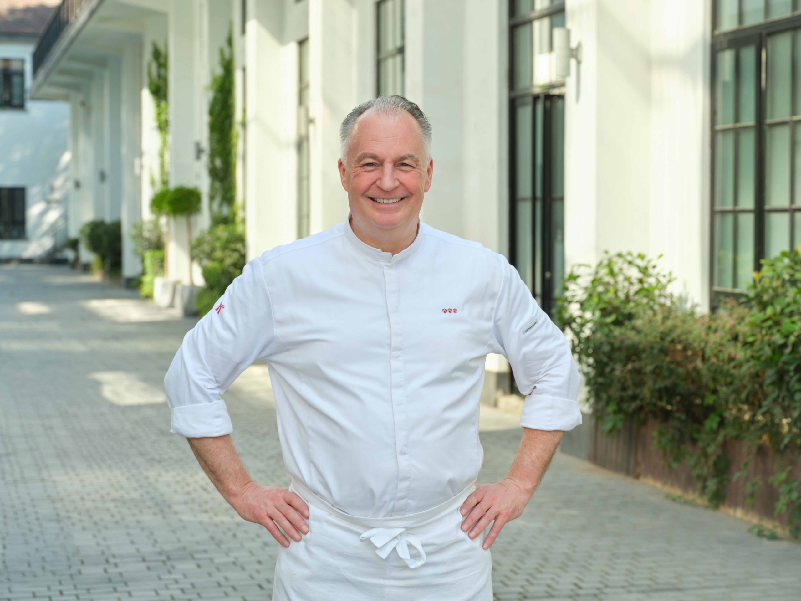 Chef Stefan Stiller landscape - ChefHeads-Magazin Smiling male chef in a white uniform standing with hands on hips outdoors near a modern building.