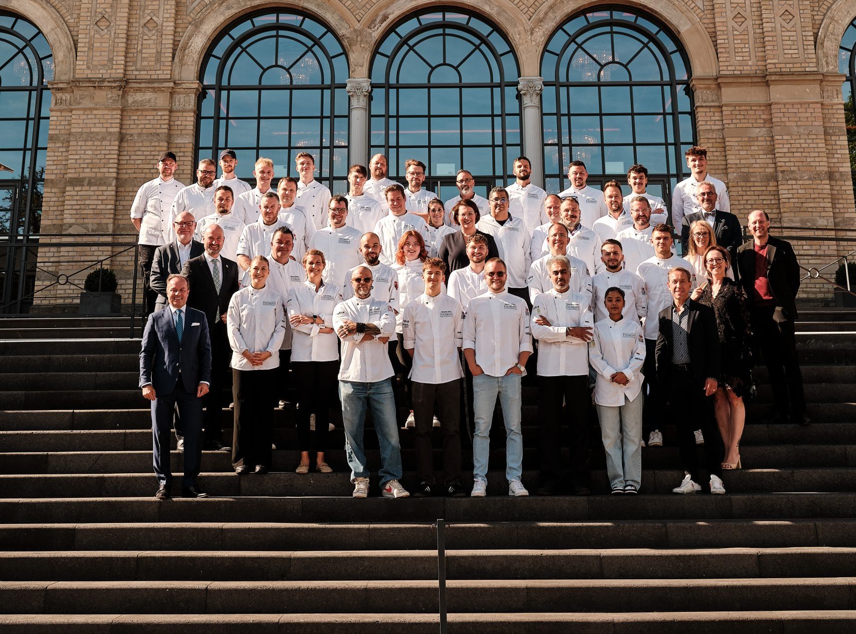 Group of chefs and staff in white uniforms pose on the steps in front of a grand brick building with large rounded, decorative windows.