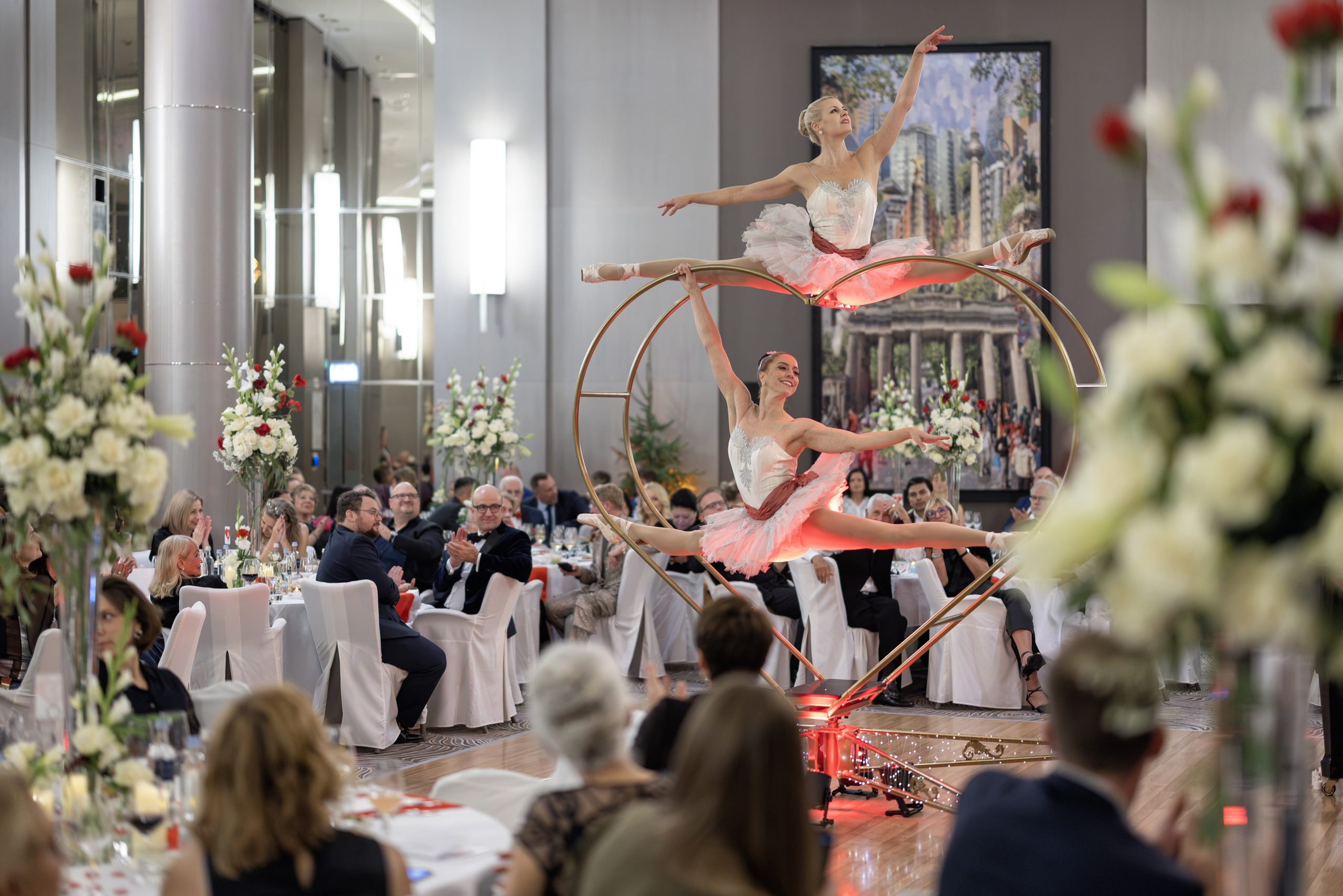 WA_Berlin_2026_G2A0147 - Aerial ballet performers in pink tutus on a Cyr wheel entertaining guests at a formal banquet hall banquet with floral decorations and an attentive audience.